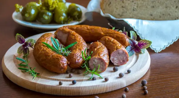 Kiełbasa podwawelska na desce z rozmarynem, obok ogórki i chleb.