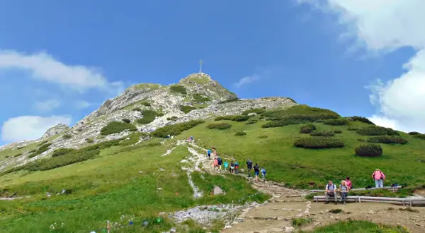 Tłum turystów wspina się kamienistym szlakiem na Giewont. Na szczycie widoczny krzyż.