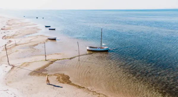 Spokojna plaża w Kuźnicy, kilka łodzi na płytkiej wodzie, jedna z masztem. Osoba w słomkowym kapeluszu spaceruje brzegiem.