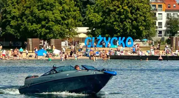 Łódź motorowa płynie po jeziorze, w tle plaża z ludźmi i napis "GIŻYCKO". Idealne miejsce, gdzie na Mazury!