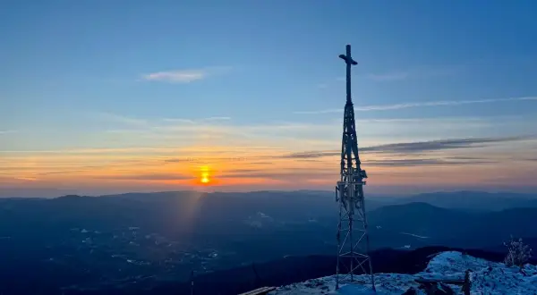 Zachód słońca nad górami, wieża z krzyżem na szczycie, ławki pokryte śniegiem. W oddali widać lasy, w tym smerek.