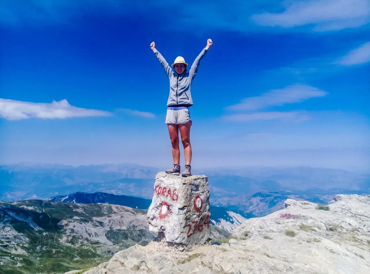 Szczyt Korab zdobyty! Radosna turystka z rękami w górze na skale z napisem "KORAB" i wysokością 2764 m.