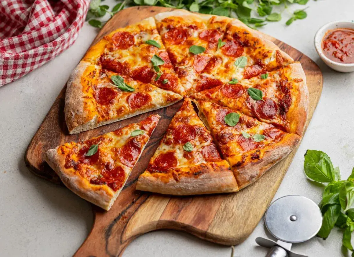 Pokrojona pizza pepperoni z serem i bazylią na desce, obok sos i nóż do pizzy.