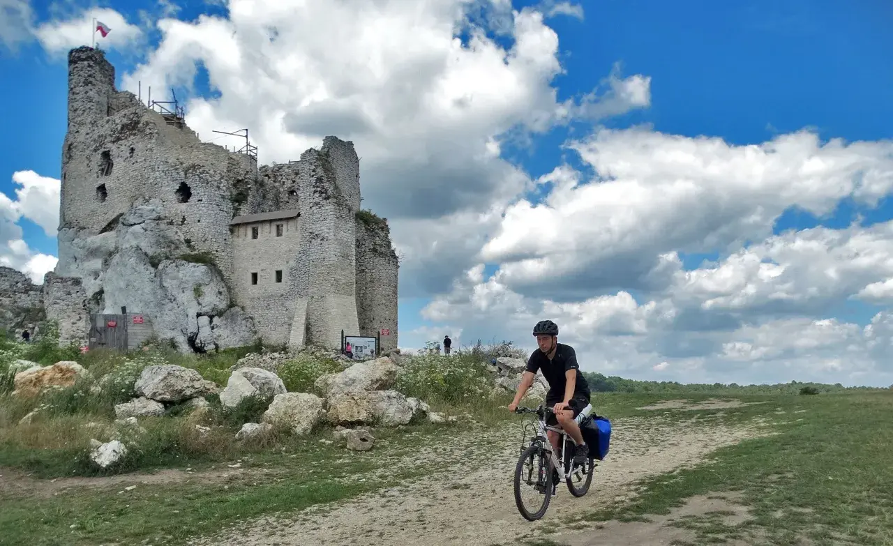 Rowerzysta przemierza malownicze **Jura Krakowsko-Częstochowska szlaki**, mijając ruiny zamku na skale.