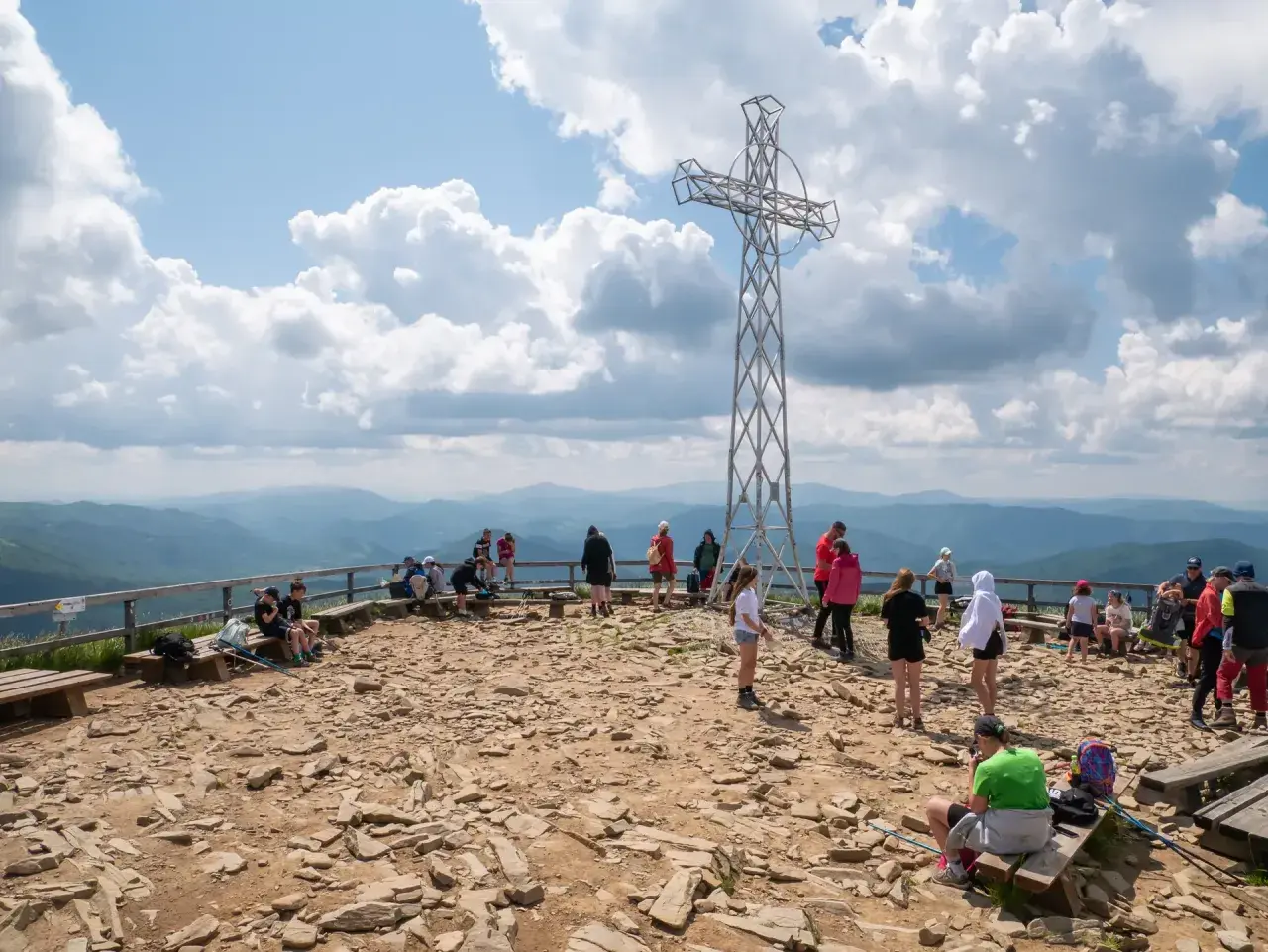 Ludzie odpoczywają na szczycie, podziwiając widoki na Bieszczady. Metalowy krzyż góruje nad kamienistym terenem, a w oddali widać łagodne pasma górskie.