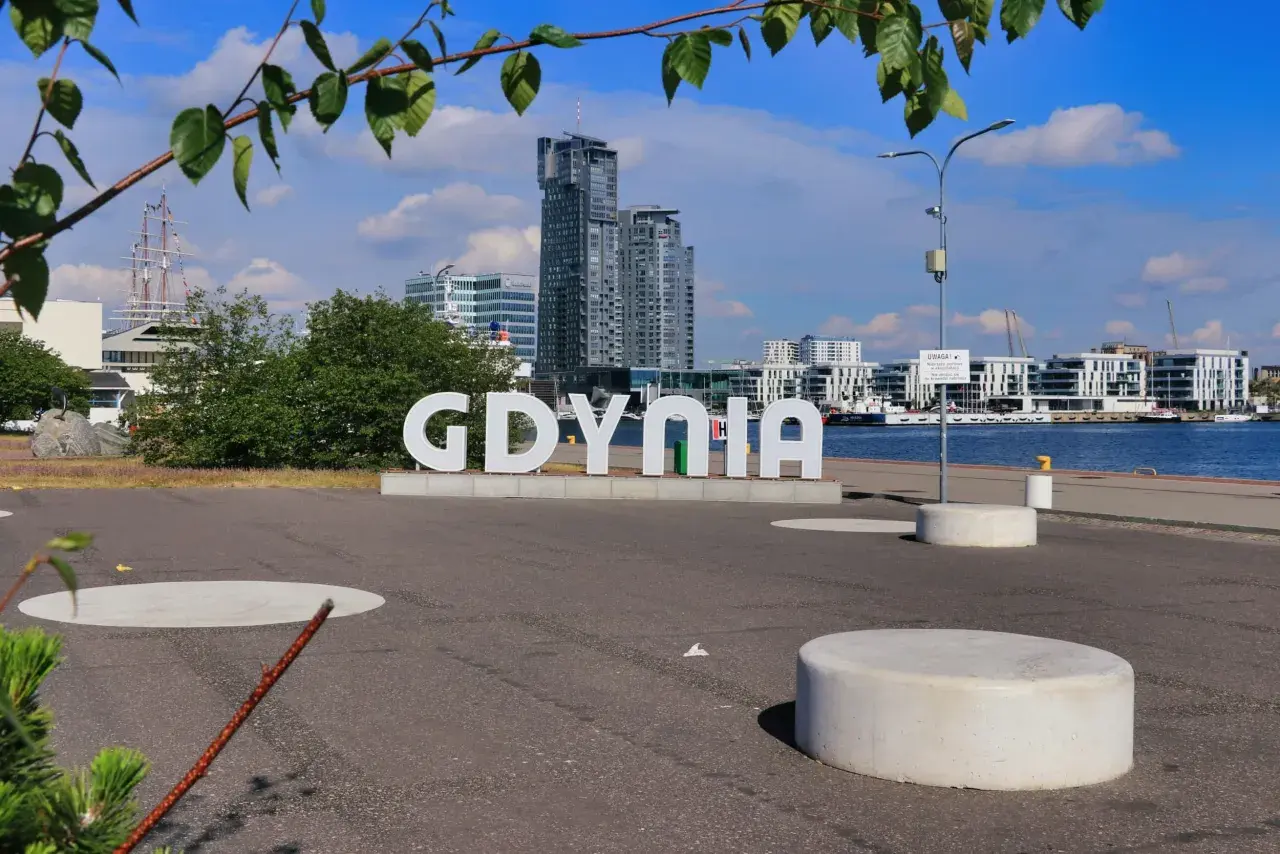Napis "GDYNIA" na tle nowoczesnych budynków i żaglowca. Widok na port, idealny na spacer po gdynia stare miasto.