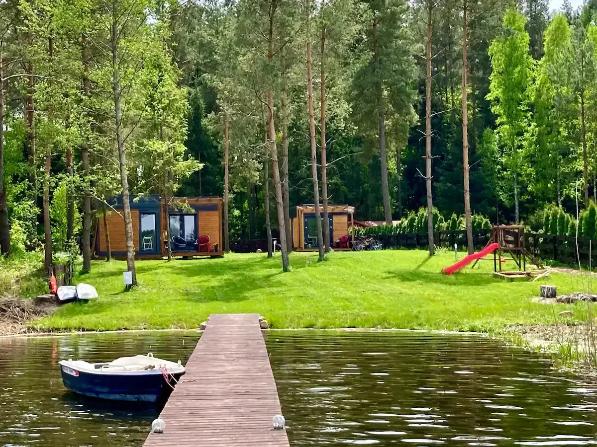 Drewniany pomost prowadzi do jeziora, gdzie cumuje łódka. Na brzegu, wśród drzew, stoją domki letniskowe, plac zabaw i rowery. Idealne miejsce na wypoczynek w **kiekrz**.
