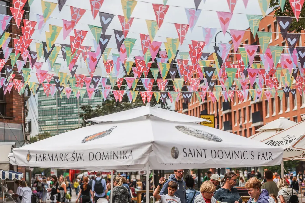 Kolorowe chorągiewki ozdabiają ulicę podczas Jarmarku Świętego Dominika. Ludzie spacerują pod białymi parasolami z napisami "Jarmark Św. Dominika" i "Saint Dominic's Fair".