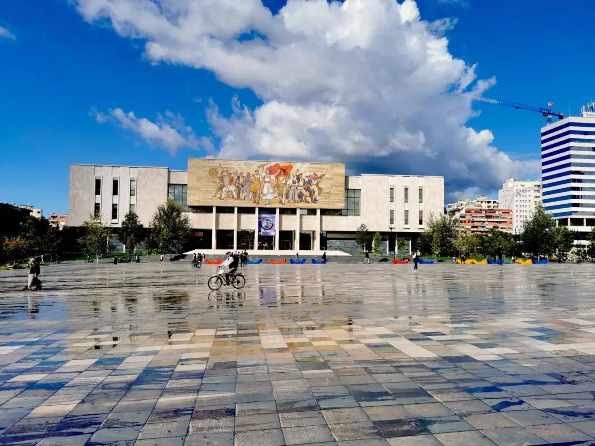 Plac w Tiranie, stolicy Albanii, z monumentalnym budynkiem i mozaiką na fasadzie. Na pierwszym planie mokra nawierzchnia odbija niebo.