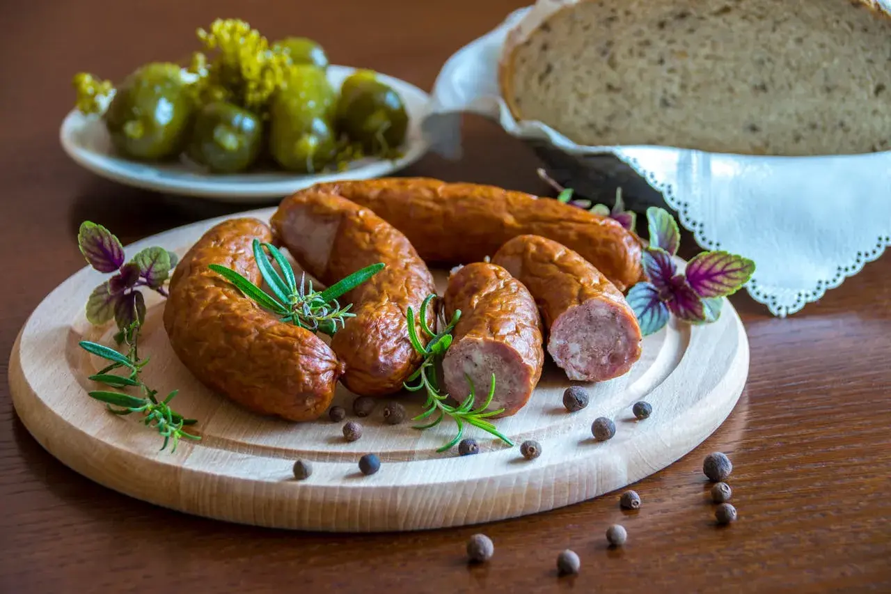 Kiełbasa podwawelska na desce z rozmarynem, obok ogórki i chleb.