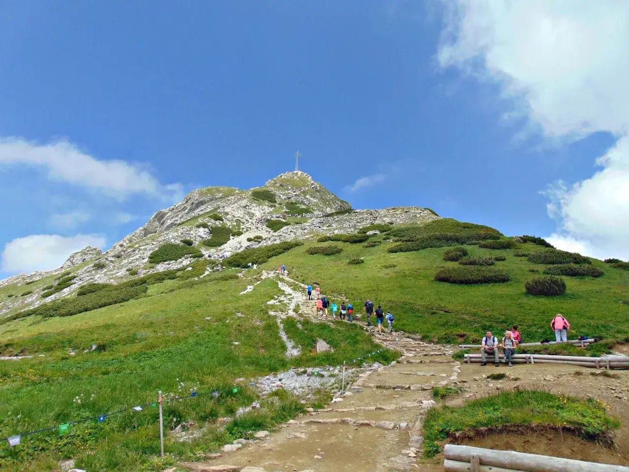 Tłum turystów wspina się kamienistym szlakiem na Giewont. Na szczycie widoczny krzyż.