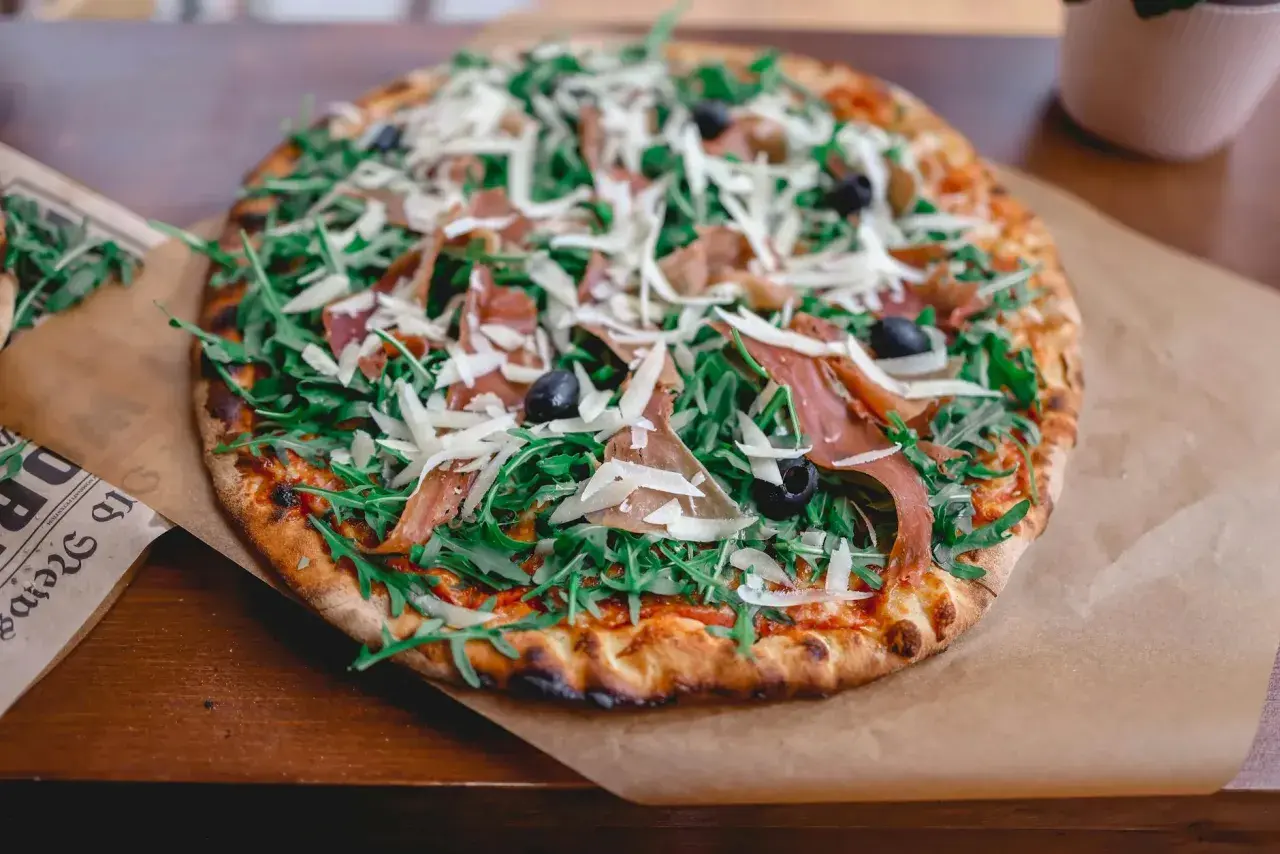 Pyszna pizza parma z rukolą, oliwkami i parmezanem, podana na papierze.