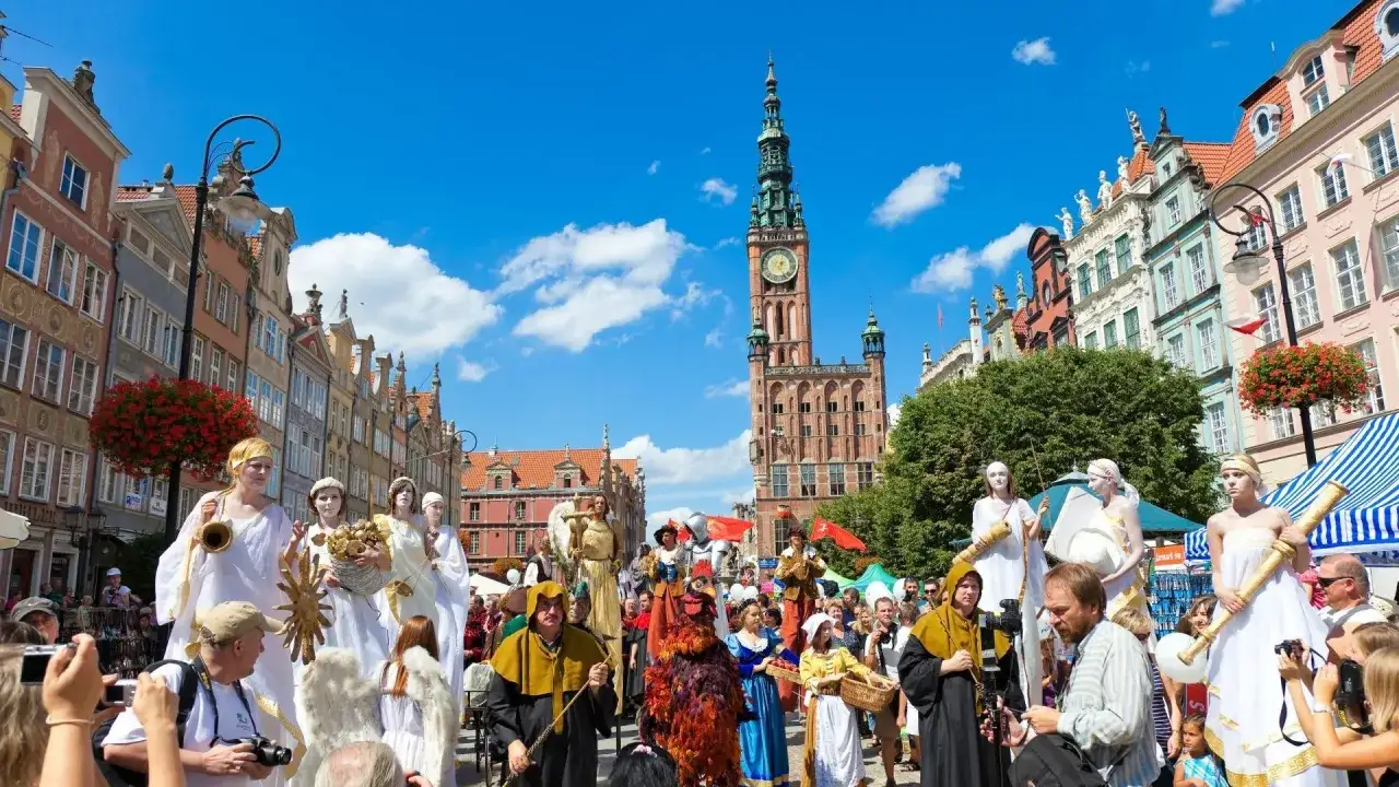 Barwny jarmark dominikański w Gdańsku. Ludzie w strojach z epoki, postaci na szczudłach i tłumy widzów na tle zabytkowych kamienic.