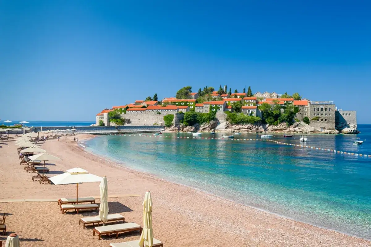 Słoneczna plaża w Czarnogórze z leżakami i parasolami, w tle wyspa Sveti Stefan z czerwonymi dachami i turkusowe morze.