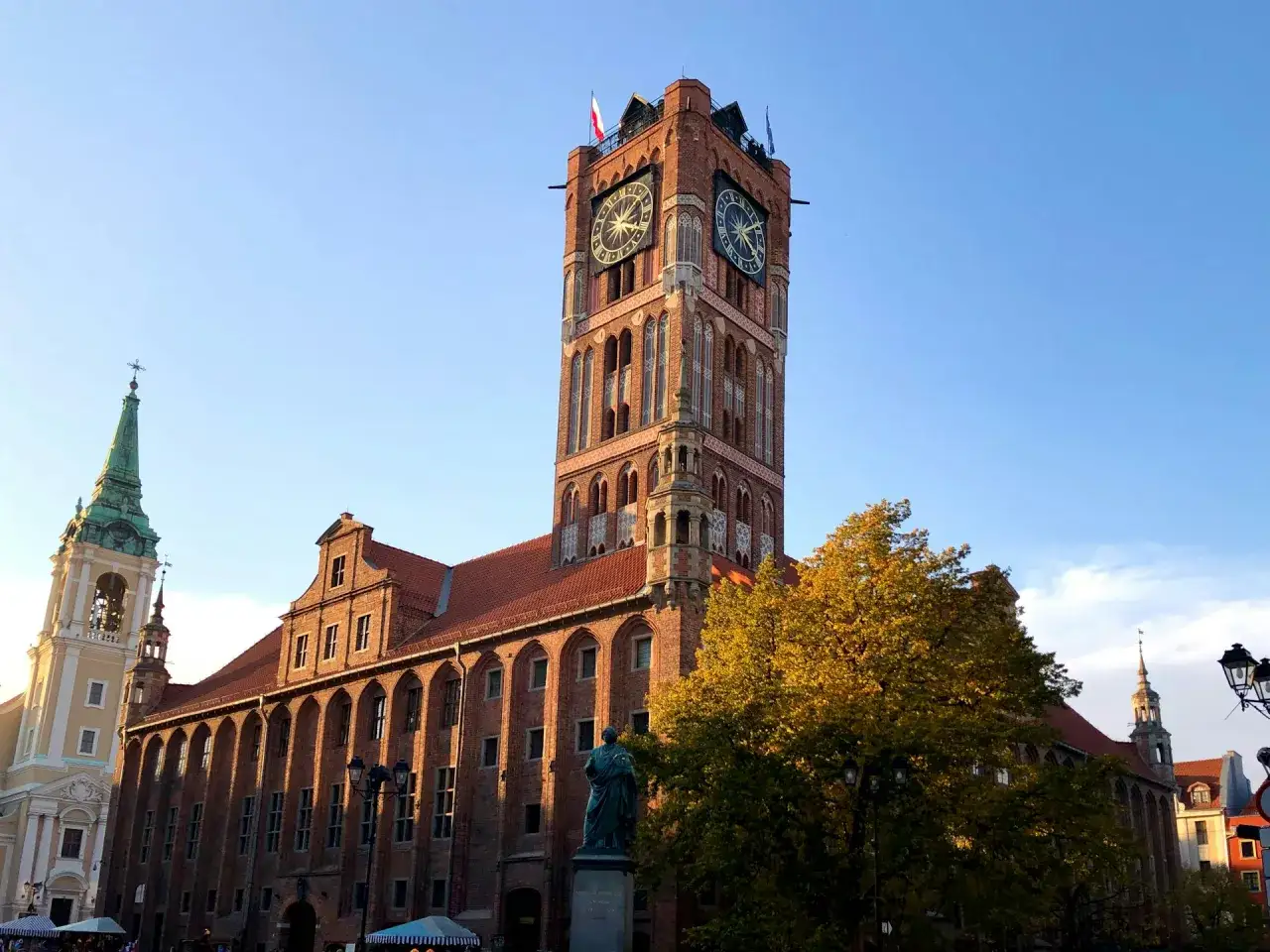 Toruń stare miasto: Ratusz Staromiejski z zegarami i wieżą, obok kościół z zieloną wieżą. Złote liście drzewa dodają uroku.