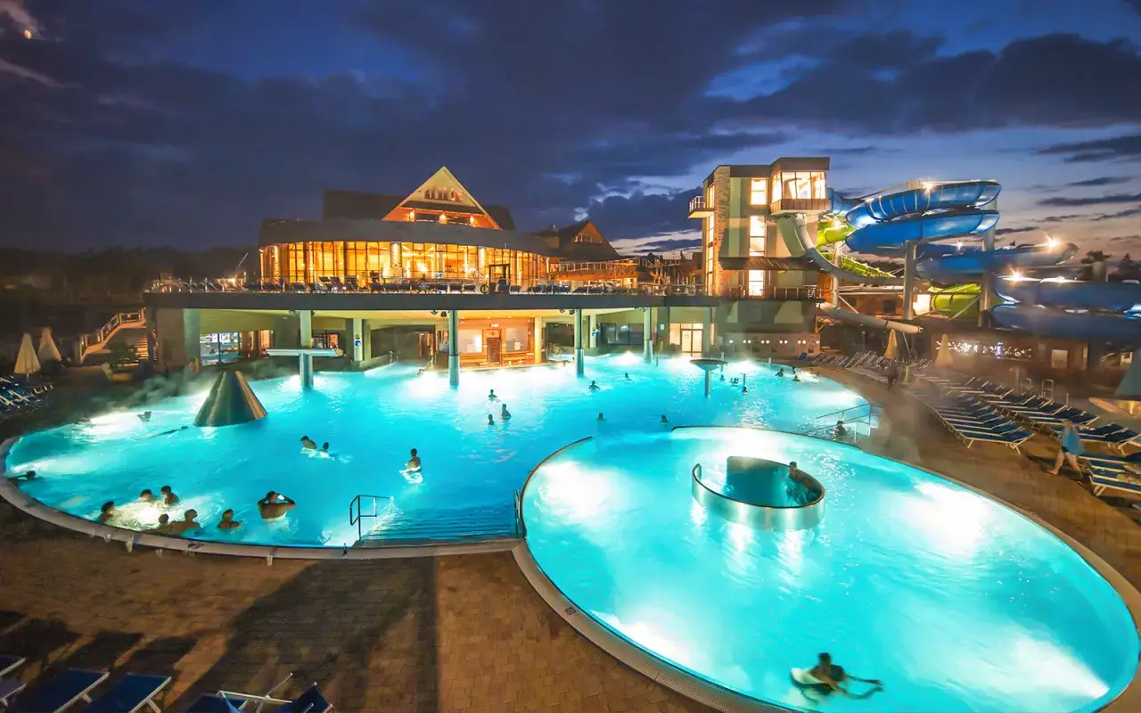 Relaks w hotelach w Zakopanem z basenem. Kąpiel w podgrzewanych wodach pod nocnym niebem, z widokiem na zjeżdżalnie i oświetlone budynki.