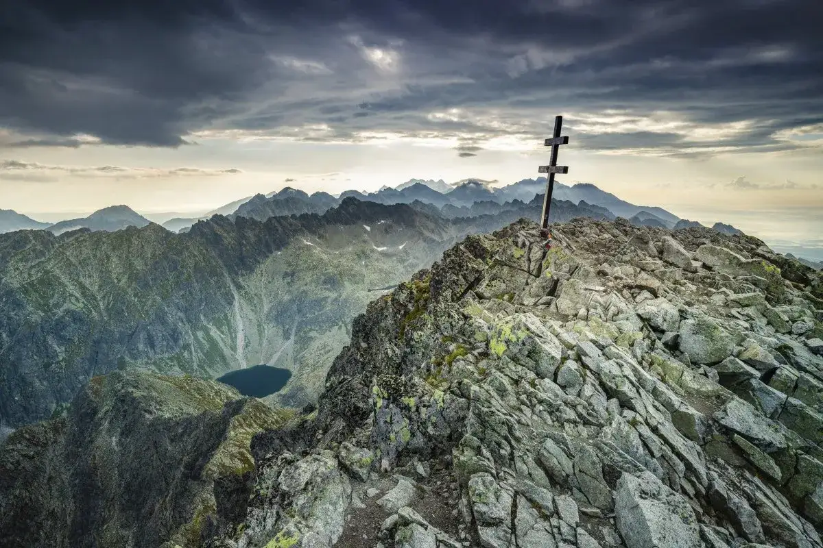 Kamienisty szczyt w Tatrach Wysokich 2494 m z krzyżem. W dole jezioro, w tle pasmo górskie pod burzowym niebem.