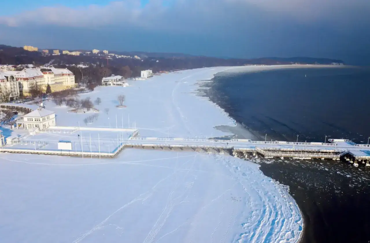 Mroźna zima w Polsce. Zamrożone morze i plaża pokryte śniegiem, z widocznym molo i budynkami w oddali.