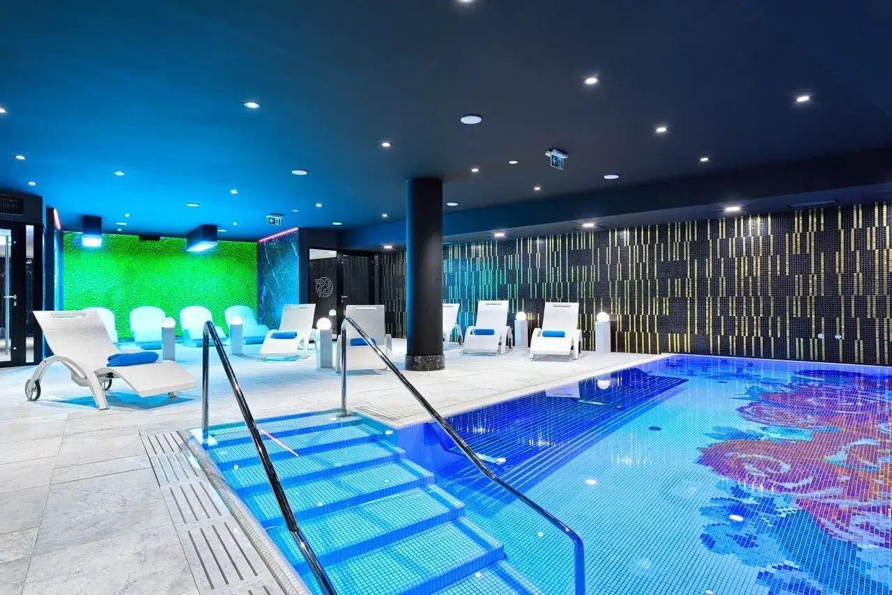 Nowoczesny basen i strefa relaksu w krakow hotel spa. Leżaki, zielona ściana i mozaika w basenie tworzą atmosferę luksusu.