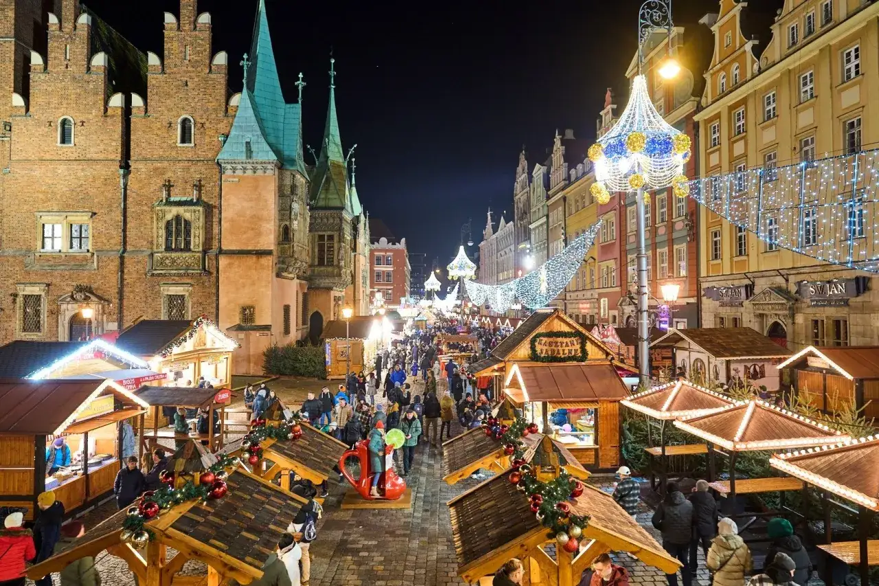 Tętniący życiem jarmark we Wrocławiu nocą, pełen ludzi, świątecznych dekoracji i drewnianych straganów.