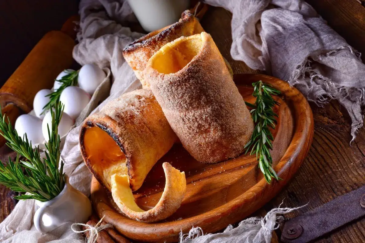 Kusząco wyglądające, posypane cukrem i cynamonem chimney cake na drewnianej desce, obok jajek i gałązki rozmarynu.