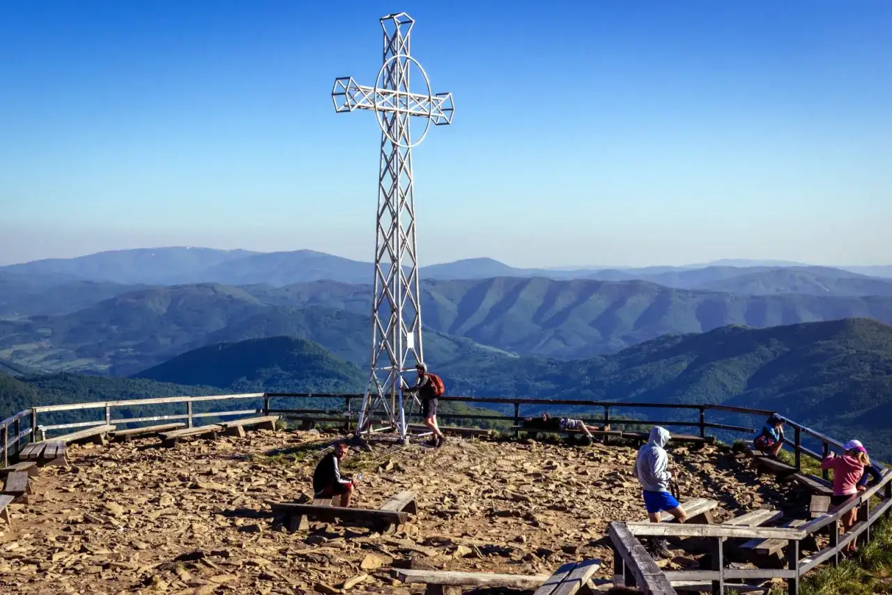 Widok na Bieszczady z krzyżem na szczycie. Turyści odpoczywają na ławkach, podziwiając malownicze szlaki górskie.