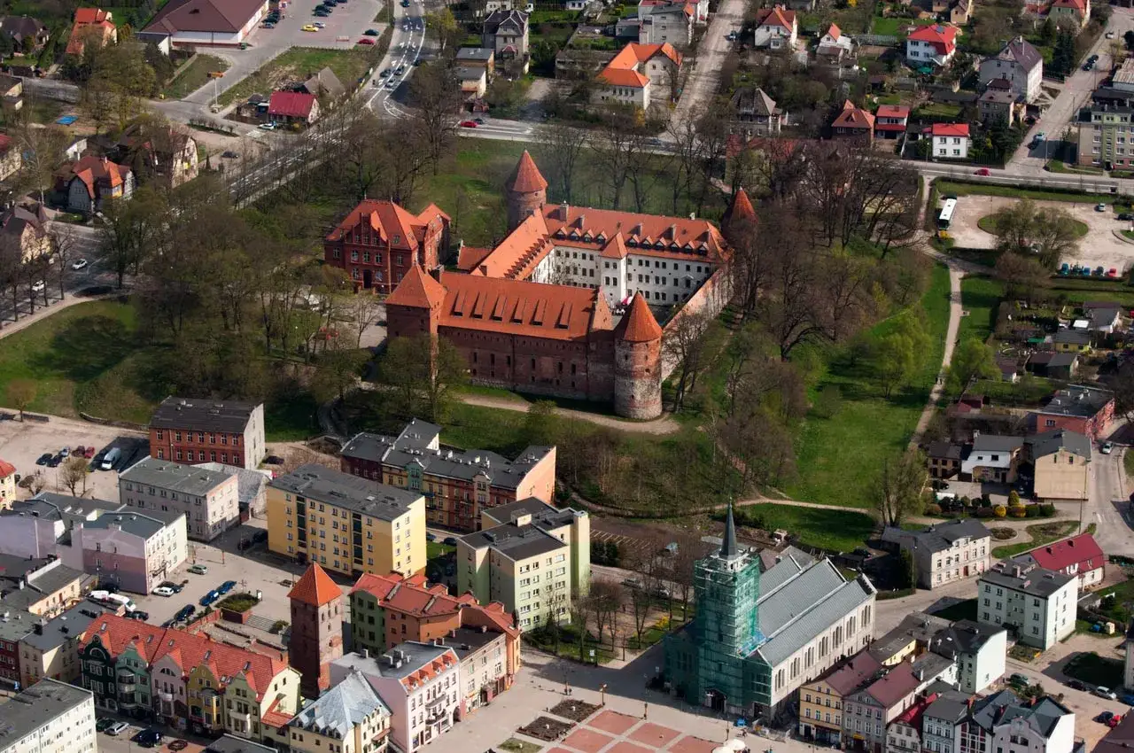 Zamek w Bytowie, rozległy kompleks z czerwonej cegły z charakterystycznymi wieżami, otoczony zielenią i zabudowaniami miejskimi.