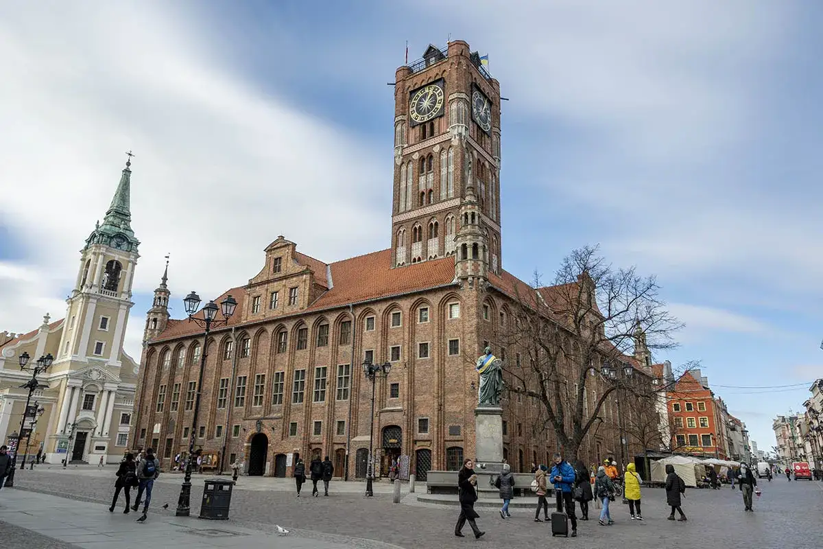 Rynek Staromiejski w Toruniu z Ratuszem i Bazyliką. Ludzie spacerują po bruku, podziwiając zabytkową architekturę.