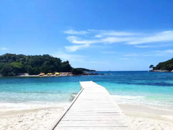 Białe molo prowadzi do krystalicznie czystej wody na Ksamil Beach. W oddali zielone wyspy i błękitne niebo.