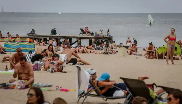 Tłum ludzi na plaży, opalanie się i relaks. Idealna pogoda na lipiec nad morzem, widać żaglówkę i wiatrak.