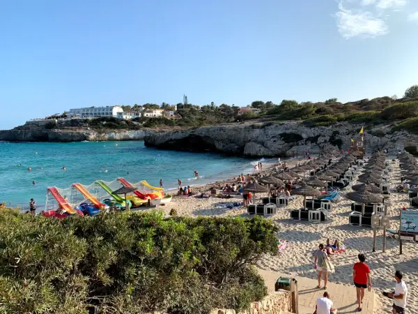 Słoneczna plaża Cala Domingos tętni życiem. Woda jest turkusowa, a na piasku widać rzędy parasoli i leżaków. Dzieci bawią się na kolorowych zjeżdżalniach.