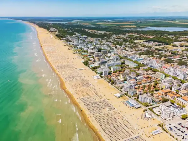 Słoneczna bibione plaża z tysiącami leżaków i parasoli, turkusowe morze i miasto w tle.
